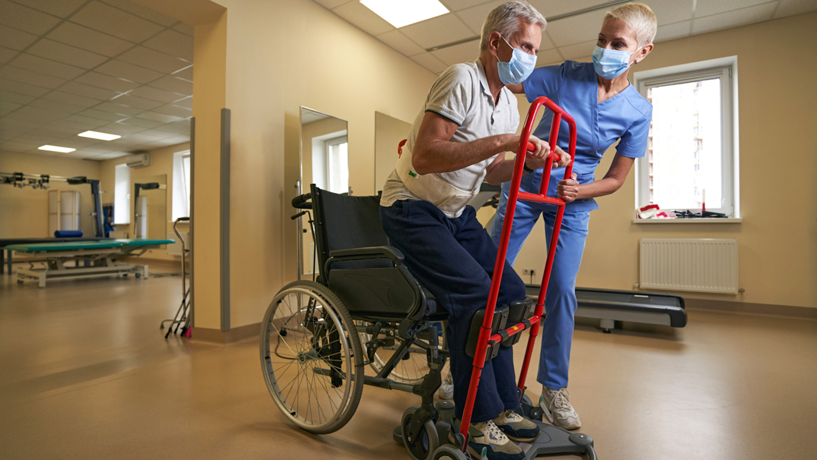 Man Being Assisted To Stand Up From Wheelchair Using An Aid And Supported By Healthcare Worker