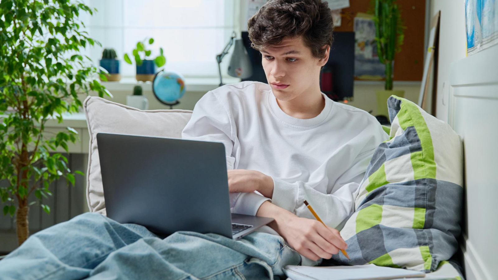Teenage boy lying on his bed while using a laptop and writing notes in a notebook.
