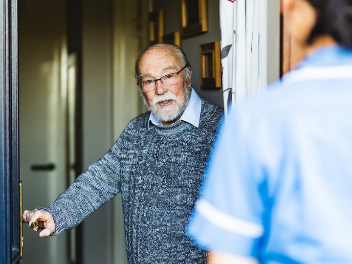 An older person stands in an open doorway speaking with a healthcare worker in a blue uniform who is visible in the foreground.