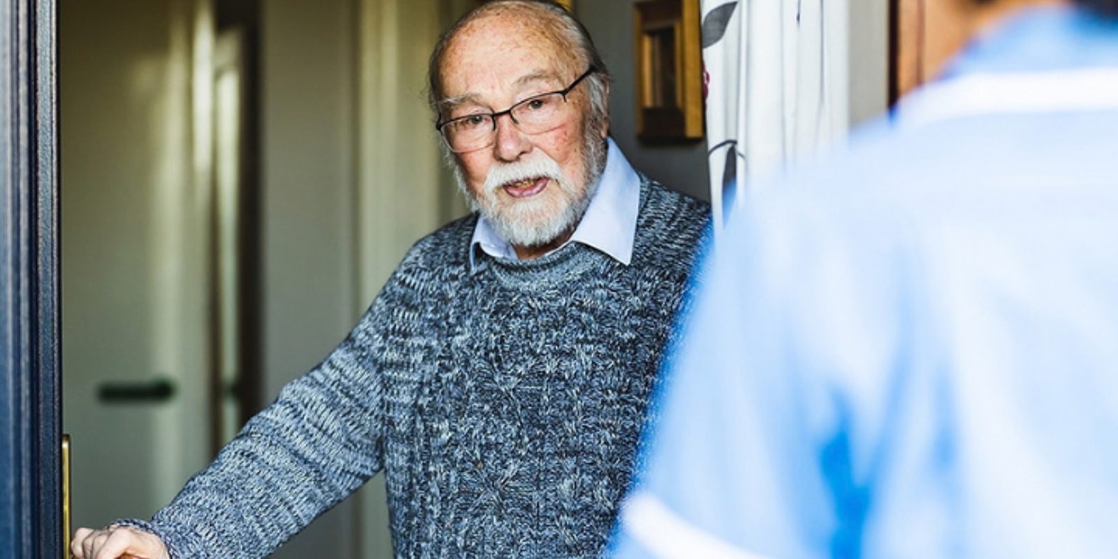 An older person stands in an open doorway speaking with a healthcare worker in a blue uniform who is visible in the foreground.