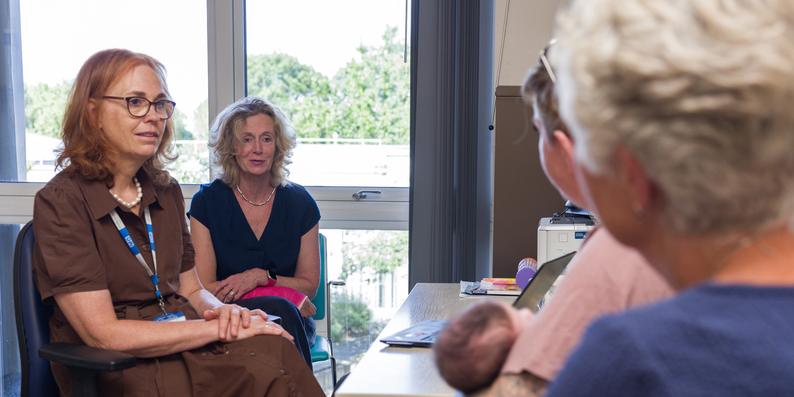 Two women sitting in a clinical discussion room, talking to a mother and another woman. The mother is holding a small baby.