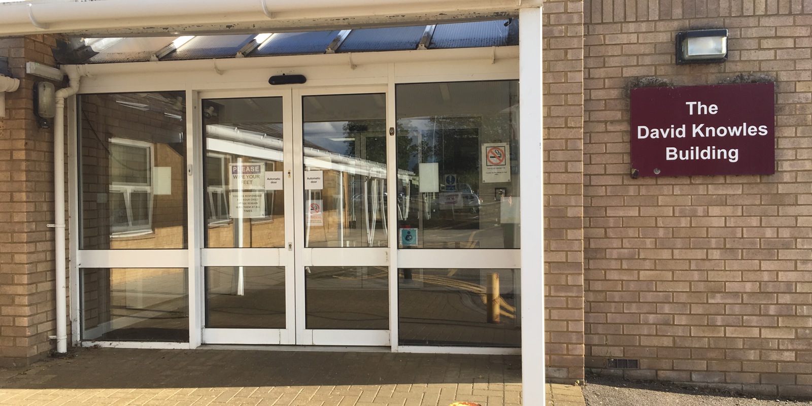 Entrance to The David Knowles Building, showing automatic glass sliding doors beneath a white covered canopy attached to a light brick building. A maroon wall sign on the right reads “The David Knowles Building.” The foreground features a paved walkway and a yellow bollard, with sunlight and blue sky reflected in the glass doors.