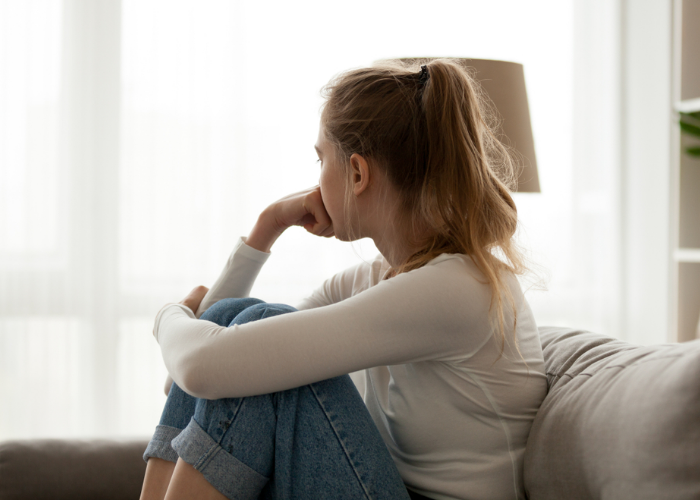Teenage girl sitting on the sofa, hugging her knees to her chest with one arm and resting her other fist on her face. She is staring out the window away from the camera.