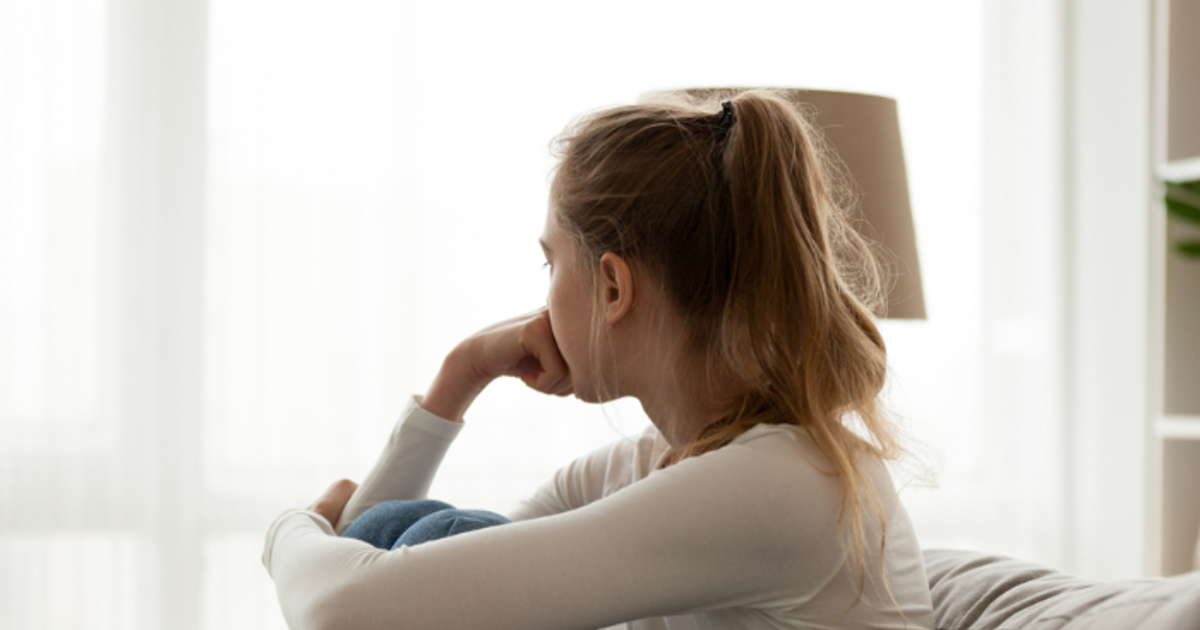 Teenage girl sitting on the sofa, hugging her knees to her chest with one arm and resting her other fist on her face. She is staring out the window away from the camera.
