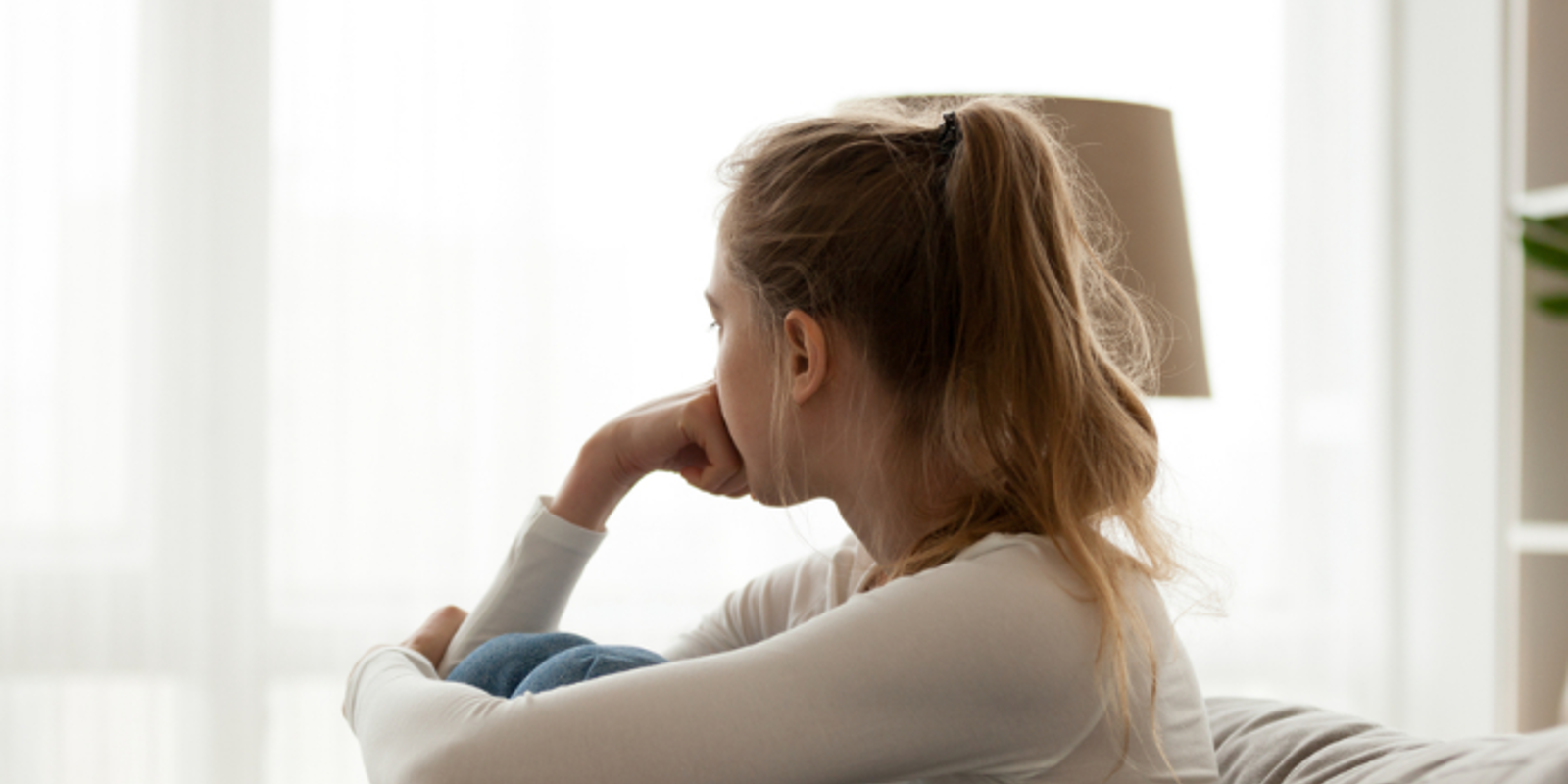 Teenage girl sitting on the sofa, hugging her knees to her chest with one arm and resting her other fist on her face. She is staring out the window away from the camera.