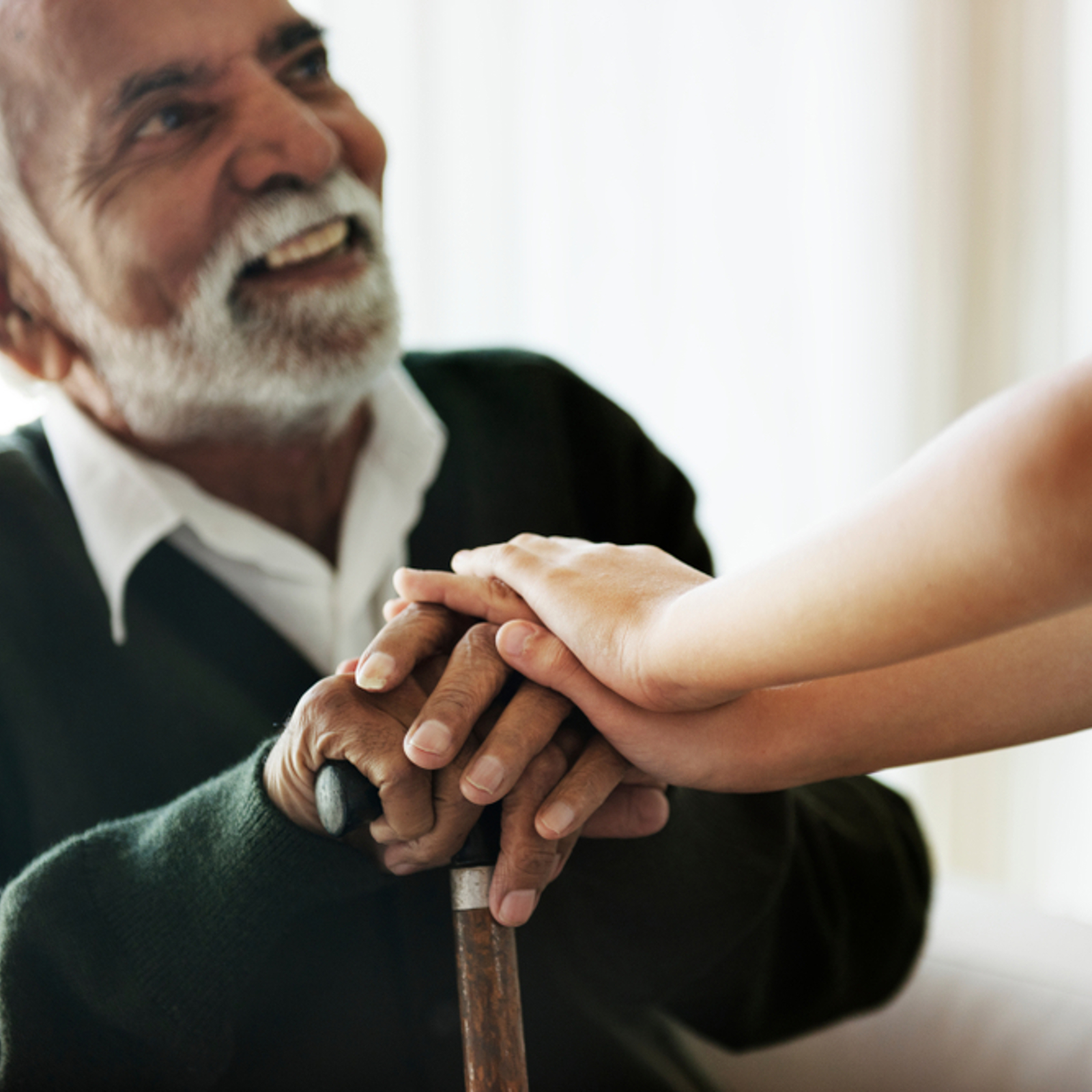An elderly man sitting and holding a walking stick. He is looking up and smiling at a child who has their hands on top of his.