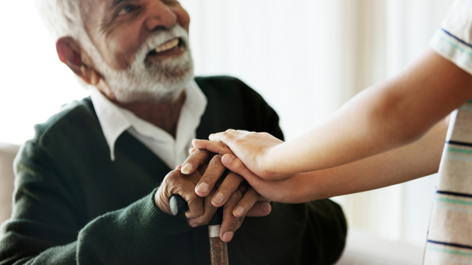 An elderly man sitting and holding a walking stick. He is looking up and smiling at a child who has their hands on top of his.