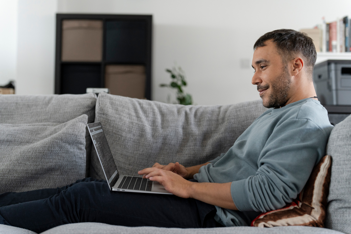 Man sitting on a sofa with his legs up and a laptop on his lap. He looking at his laptop and smiling.