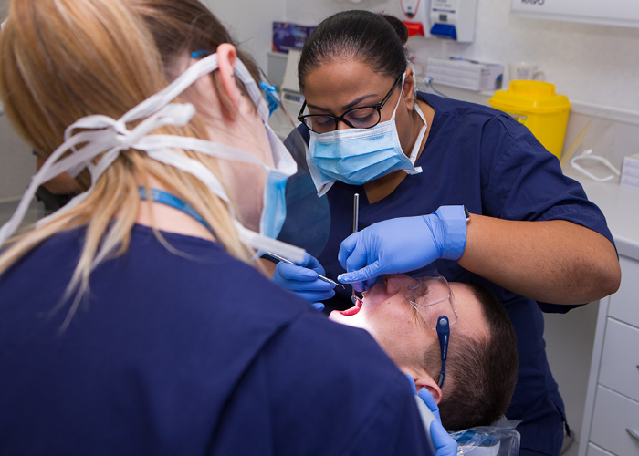 Black female dentist performing dentistry on a white male who is lying down, watched by a white female assistant.