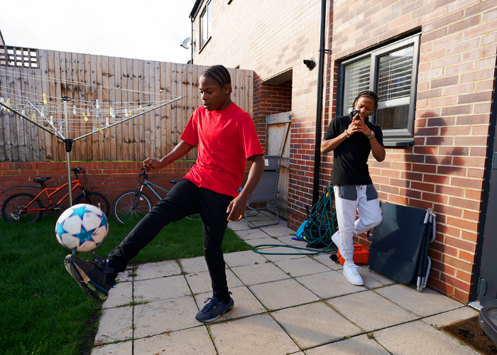 Teenage Boy Playing Kicking Ball In Back Garden With Friend Filming On Phone