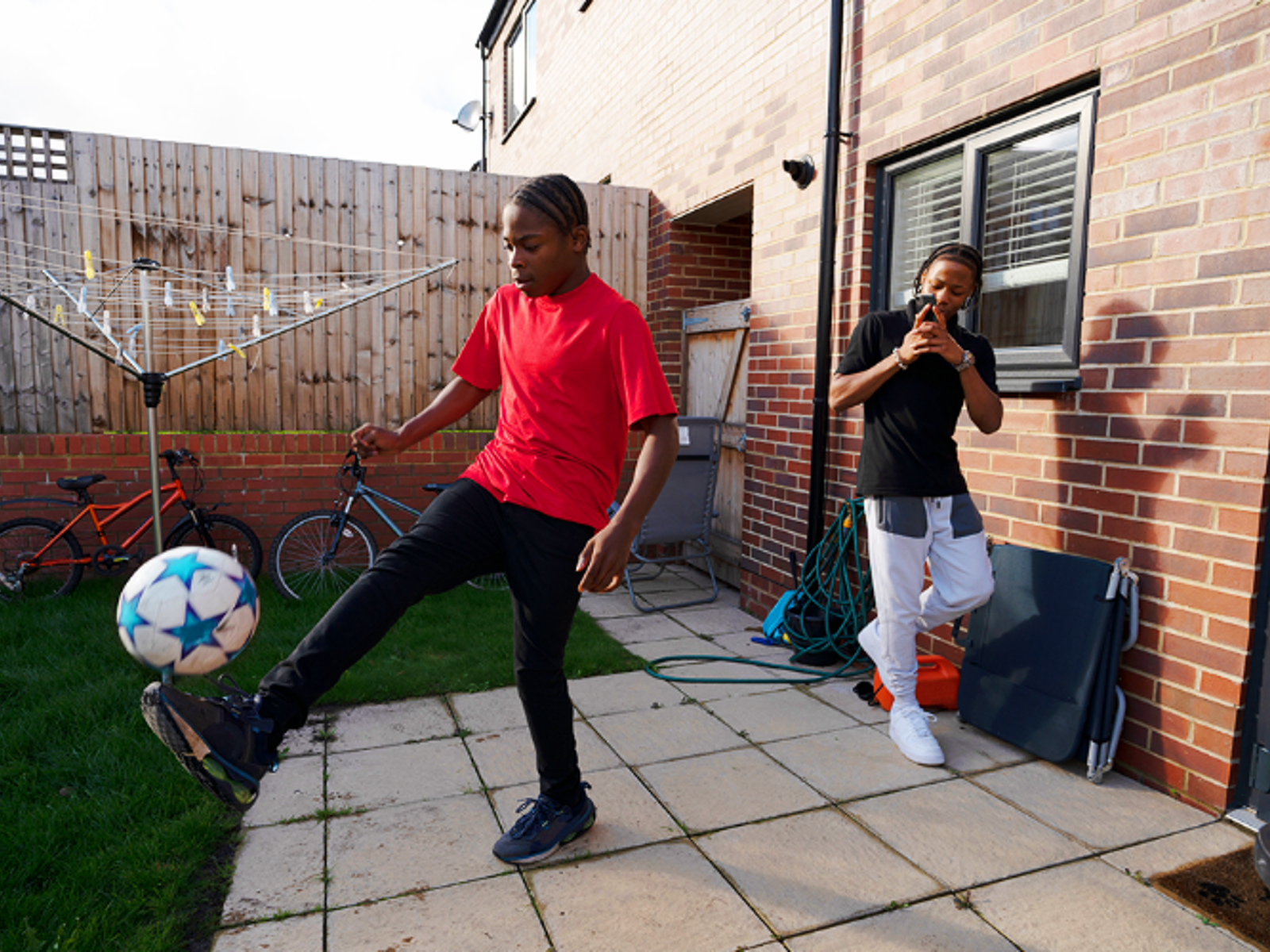 Teenage Boy Playing Kicking Ball In Back Garden With Friend Filming On Phone