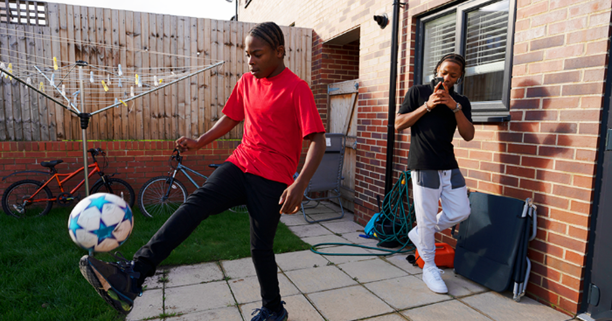 Teenage Boy Playing Kicking Ball In Back Garden With Friend Filming On Phone