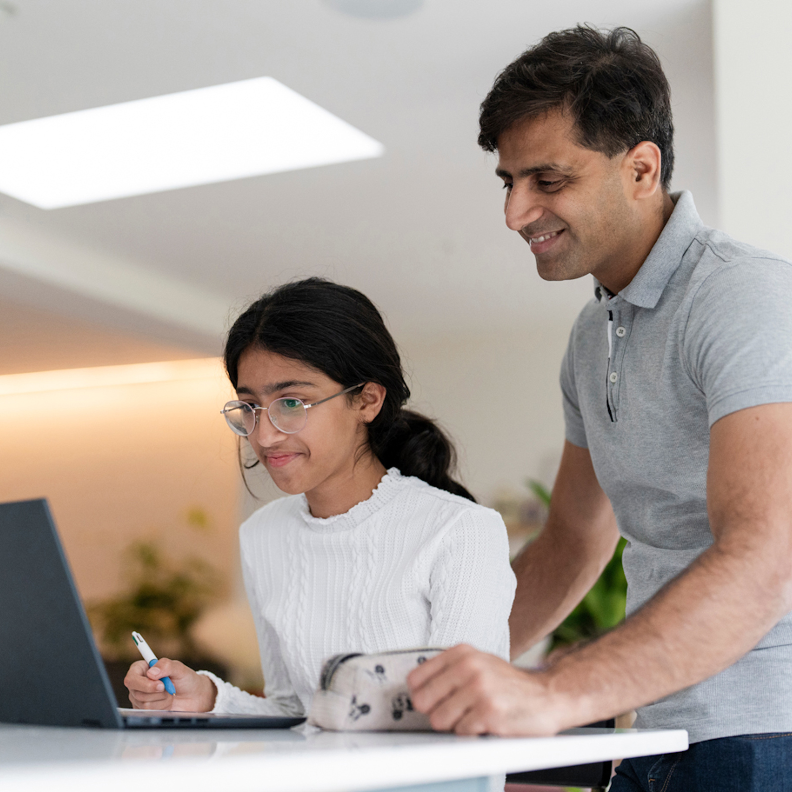 Teenage girl sitting at a table with a laptop. An adult is supervising and smiling.