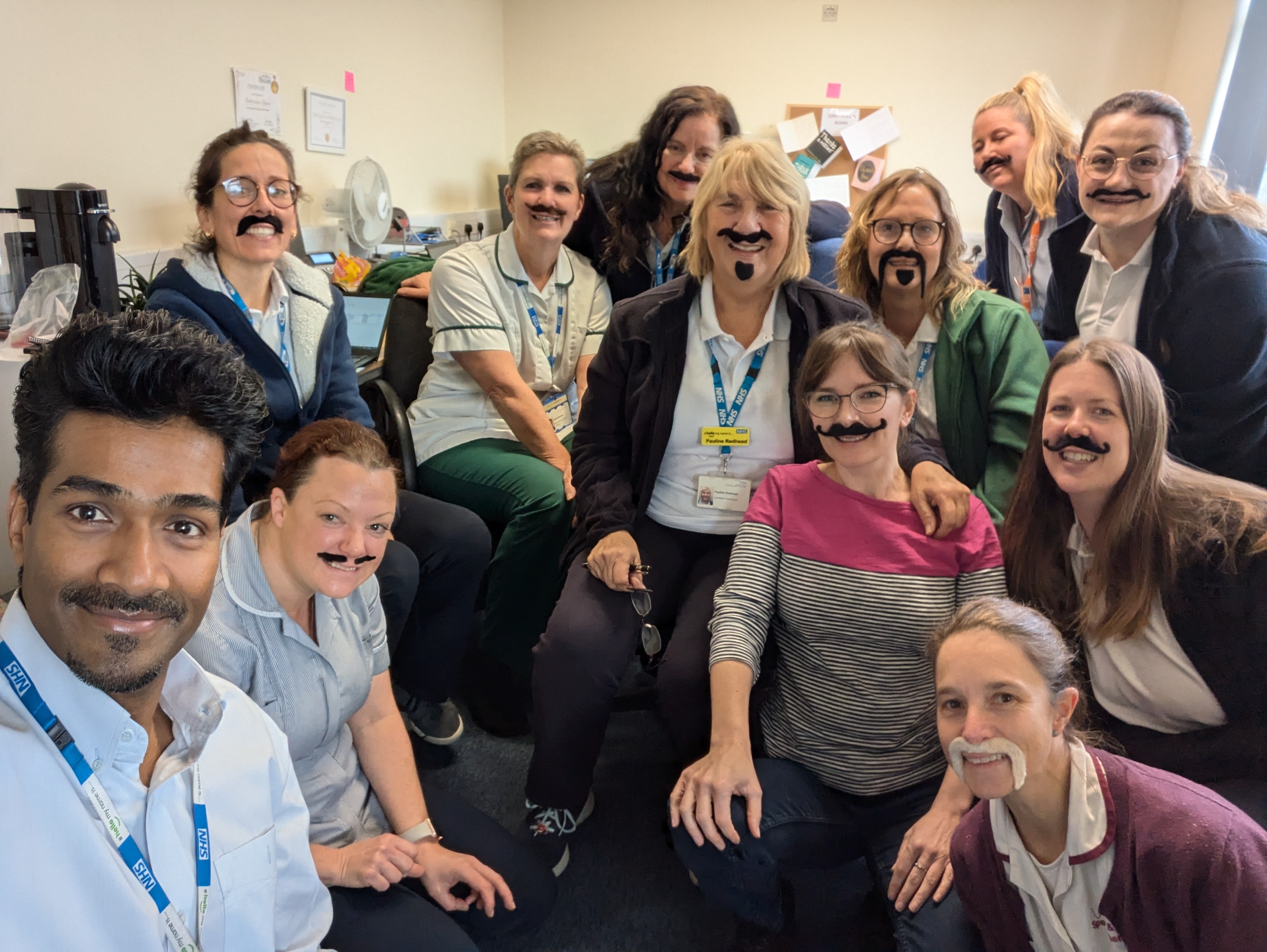 A photo of a group of NHS workers all wearing fake moustaches and smiling