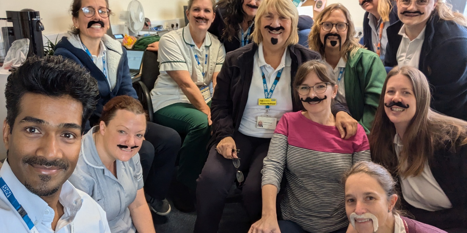 A photo of a group of NHS workers all wearing fake moustaches and smiling