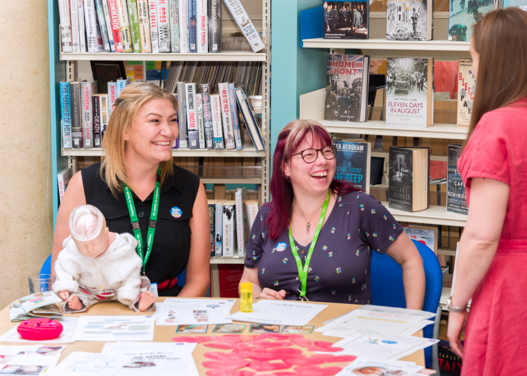 2 staff members in a library sitting at a table. They are laughing and smiling looking up at another colleague.