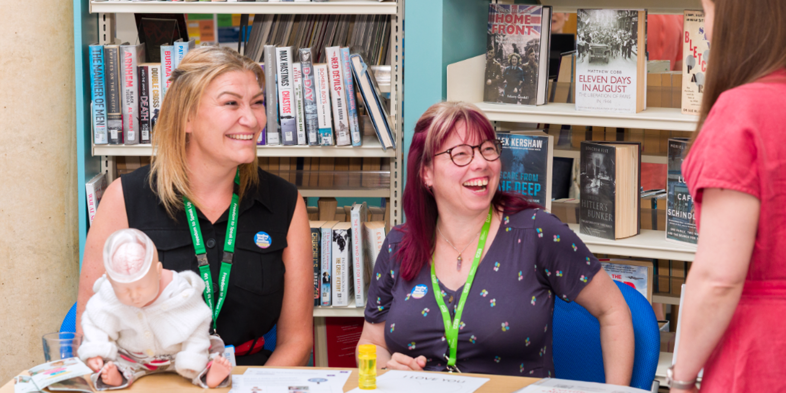 2 staff members in a library sitting at a table. They are laughing and smiling looking up at another colleague.