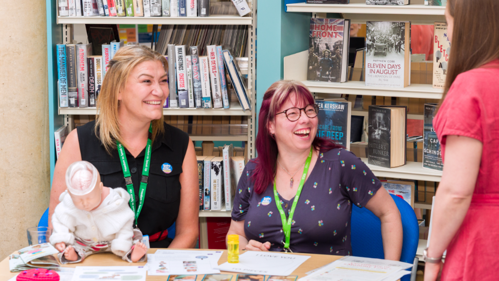 2 staff members in a library sitting at a table. They are laughing and smiling looking up at another colleague.
