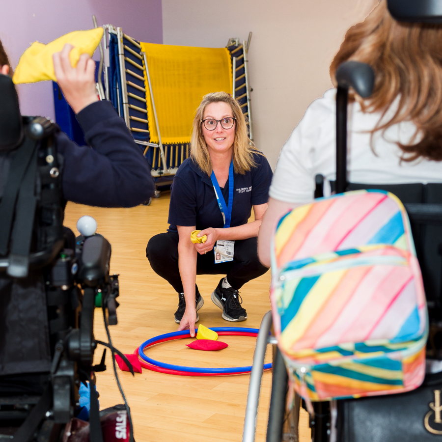 Clinician Smiling And Playing Beanbag Catch With Two Disabled Girls In Wheelchairs.