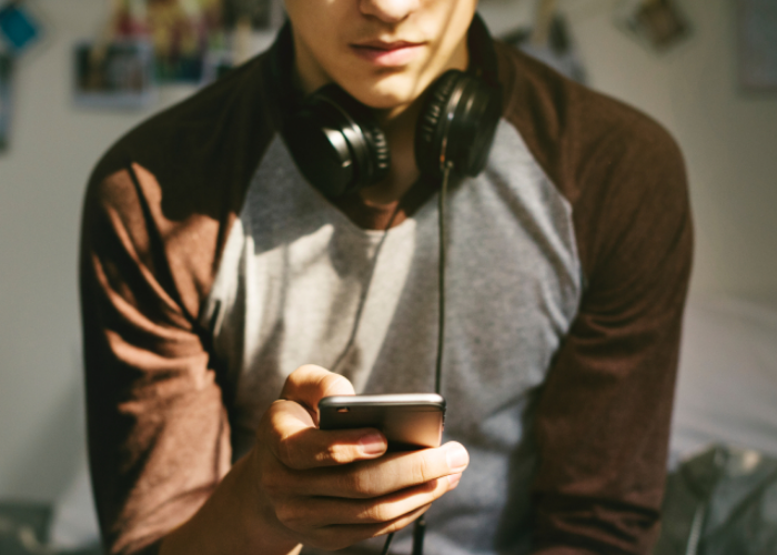 Teenage boy with headphones round his neck sitting on his bed whilst on his phone