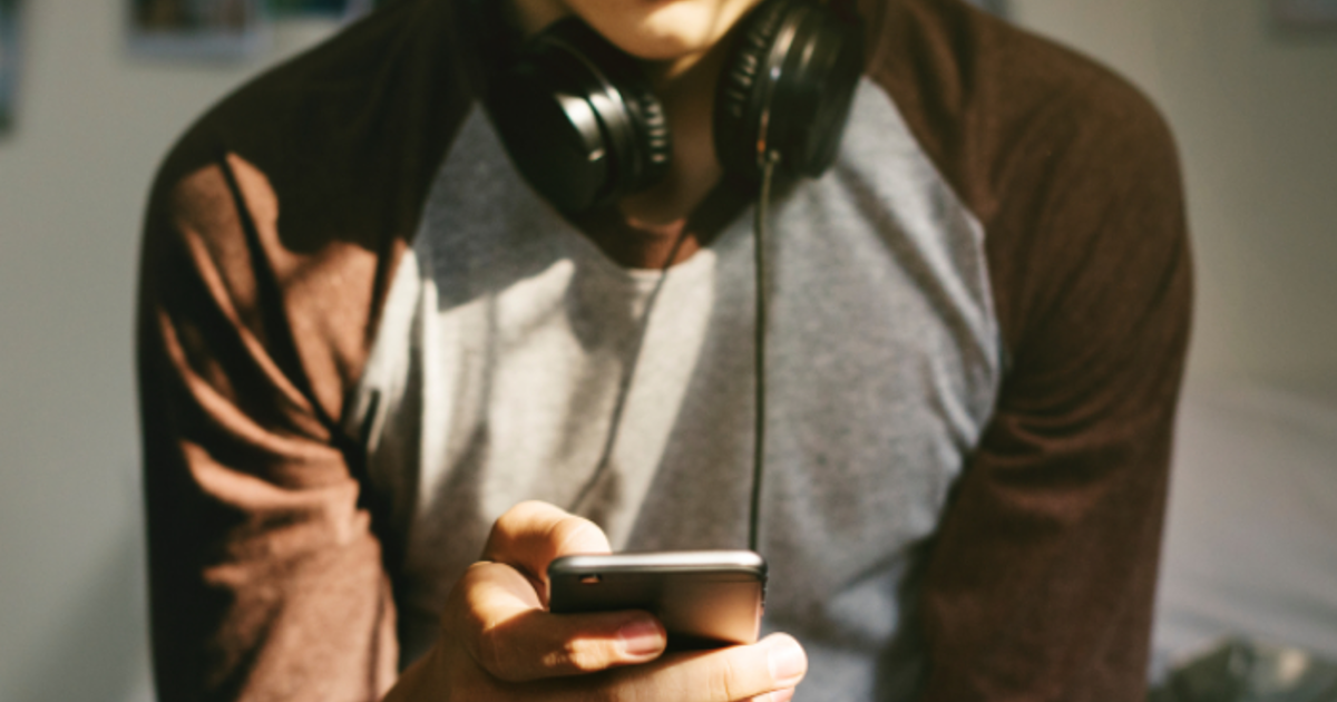 Teenage boy with headphones round his neck sitting on his bed whilst on his phone