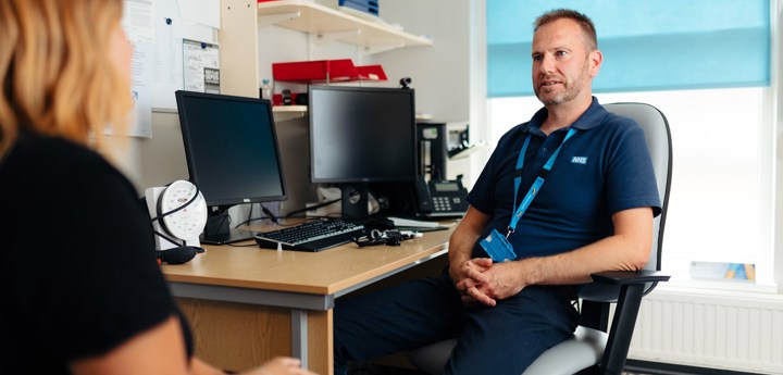 GP sitting in chair at a desk, speaking to a patient