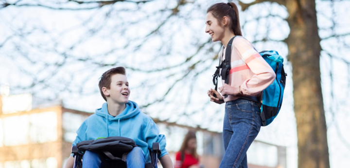 Teenage girl with backpack standing next to a teenage boy in a wheelchair outside a school