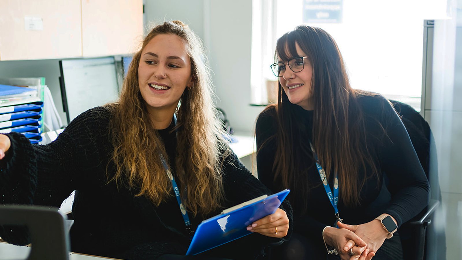 2 women in an office looking at a screen and smiling.