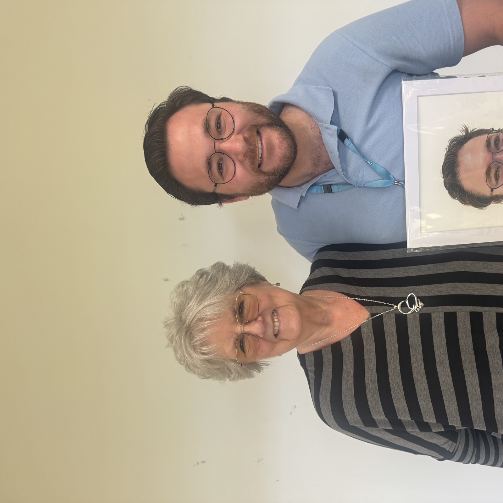 A smiling man wearing an NHS lanyard standing with a smiling older woman who is wearing a grey and black striped top. The man is holding a painted portrait of himself. 