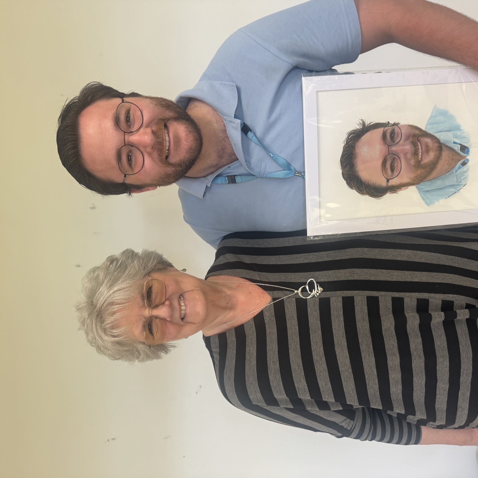 A smiling man wearing an NHS lanyard standing with a smiling older woman who is wearing a grey and black striped top. The man is holding a painted portrait of himself. 