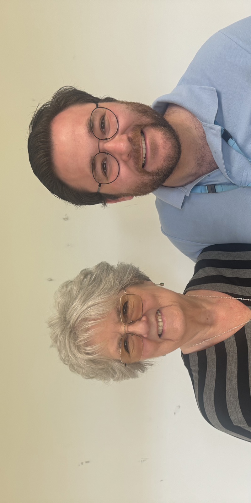 A smiling man wearing an NHS lanyard standing with a smiling older woman who is wearing a grey and black striped top. The man is holding a painted portrait of himself. 