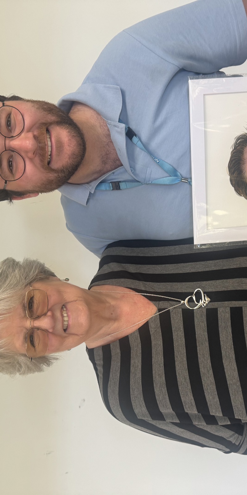 A smiling man wearing an NHS lanyard standing with a smiling older woman who is wearing a grey and black striped top. The man is holding a painted portrait of himself. 