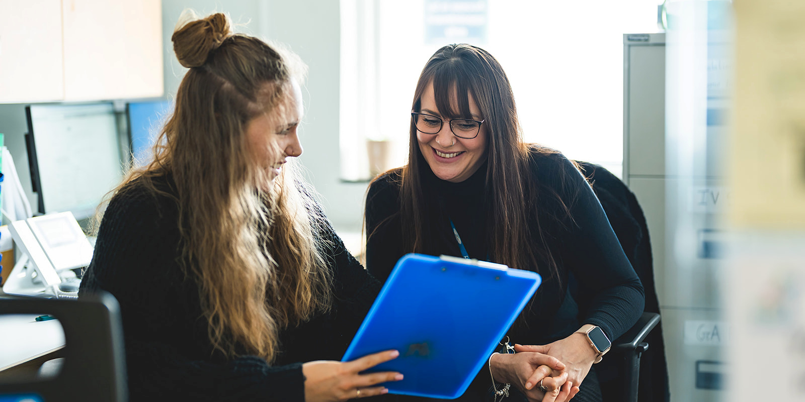 2 women in an office smiling and looking at a clipboard